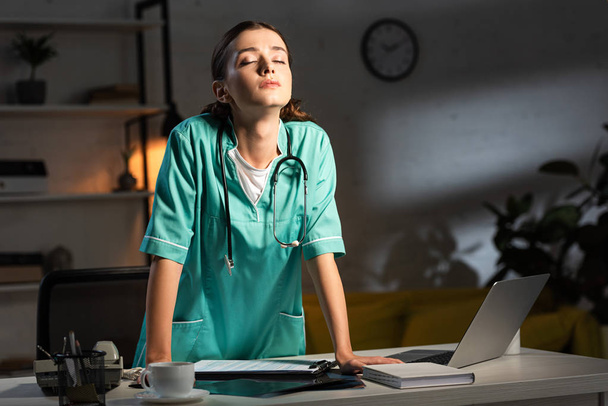 attraktive Krankenschwester in Uniform mit geschlossenen Augen während der Nachtschicht - Foto, Bild