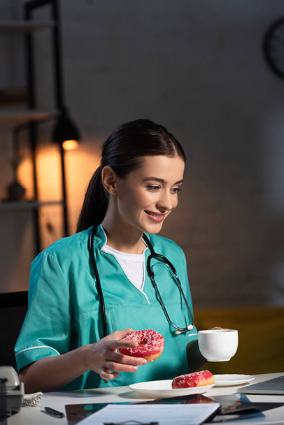 glimlachende verpleegster in uniform met donut en beker tijdens de nachtdienst - Foto, afbeelding