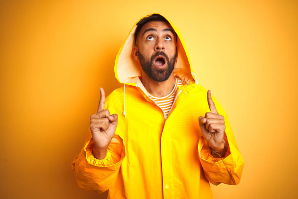 Young indian man wearing raincoat with hood standing over isolated yellow background amazed and surprised looking up and pointing with fingers and raised arms. - Fotoğraf, Görsel