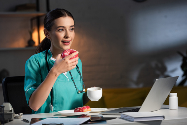 attractive nurse in uniform sitting at table and eating donut during night shift - Foto, Imagem