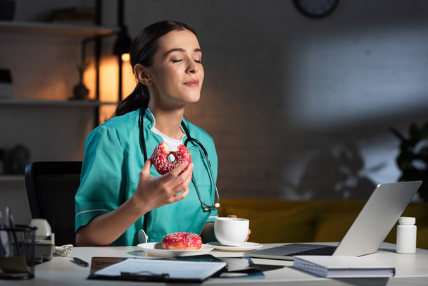 aantrekkelijke verpleegster in uniform zitten aan tafel en donut eten tijdens de nachtdienst - Foto, afbeelding
