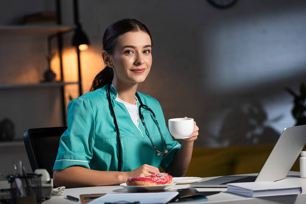 enfermeira sorrindo de uniforme sentado à mesa e segurando copo durante o turno da noite
 - Foto, Imagem