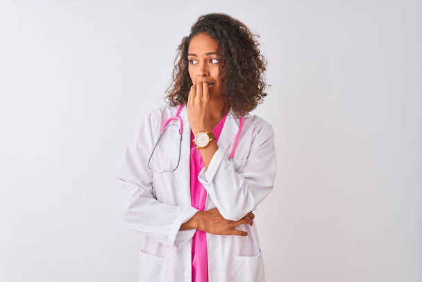 Młoda brazylijska lekarka nosząca stetoskop stojąca nad odizolowanym białym tłem wyglądająca na zestresowaną i zdenerwowaną dłońmi obgryzającymi paznokcie. Problem lękowy. - Zdjęcie, obraz