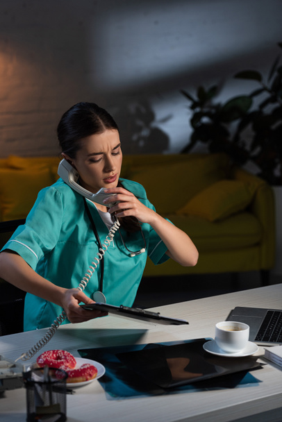 infirmière en uniforme parlant au téléphone et tenant le presse-papiers pendant le quart de nuit
 - Photo, image