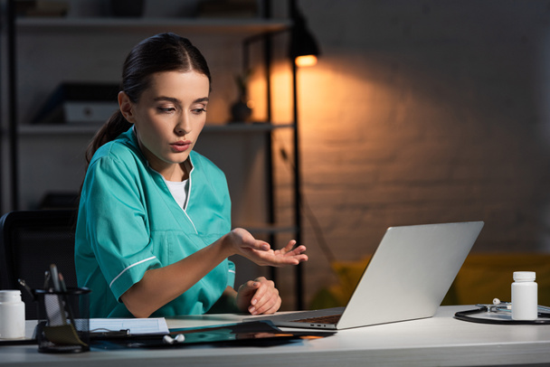 attractive nurse in uniform sitting at table and having video chat during night shift - Photo, Image
