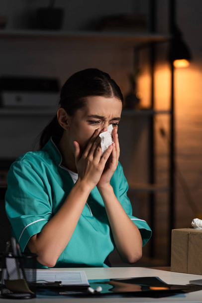 attractive nurse in uniform sneezing and holding napkin during night shift - Photo, Image