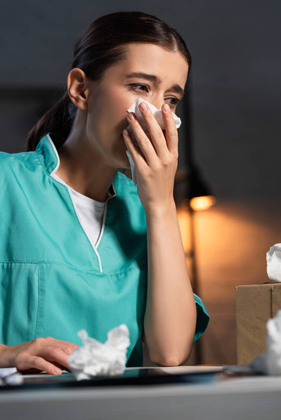atraktivní zdravotní sestra v uniformě kýchání a držení ubrousek během noční směny - Fotografie, Obrázek