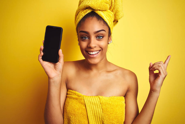 Afro mulher vestindo toalha após o chuveiro mostrando smatrphone sobre fundo amarelo isolado muito feliz apontando com a mão e o dedo para o lado
 - Foto, Imagem