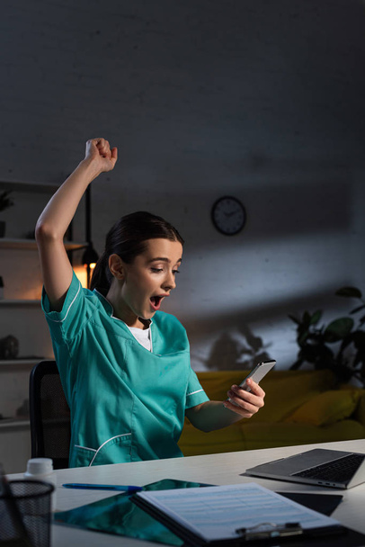 geschokte verpleegster in uniform met ja gebaar en smartphone tijdens de nachtdienst - Foto, afbeelding