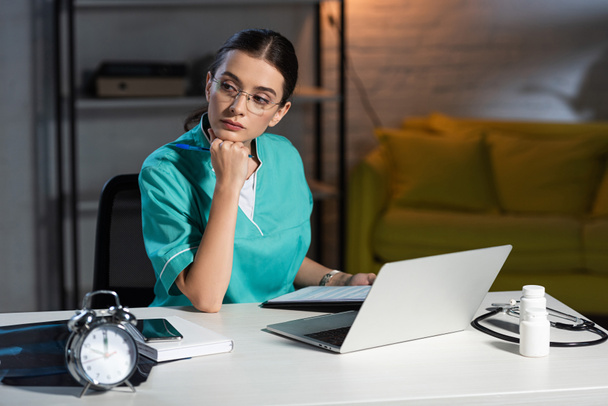 attractive nurse in uniform and glasses sitting at table during night shift - Photo, Image