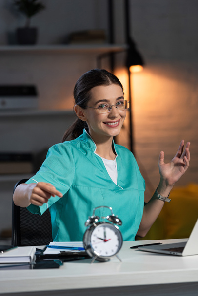 χαμογελαστή νοσοκόμα με στολή και γυαλιά που κάθεται στο τραπέζι και δείχνει με το δάχτυλο το ξυπνητήρι κατά τη διάρκεια της νυχτερινής βάρδιας - Φωτογραφία, εικόνα