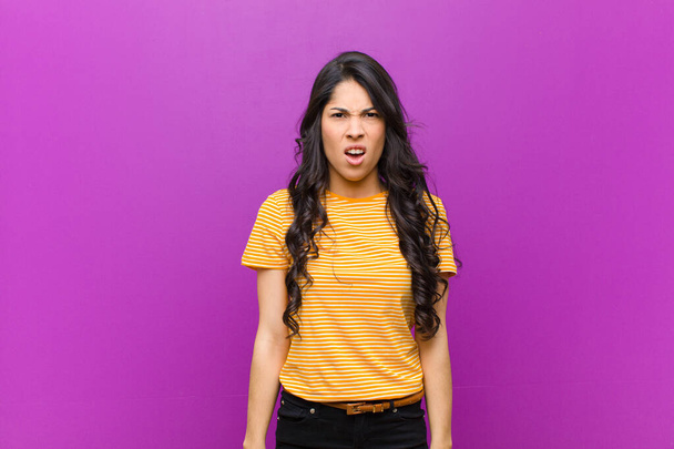 young pretty latin woman looking shocked, angry, annoyed or disappointed, open mouthed and furious against purple wall - Foto, Bild