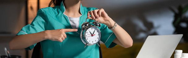 panoramic shot of nurse in uniform pointing with finger at alarm watch during night shift - Fotoğraf, Görsel