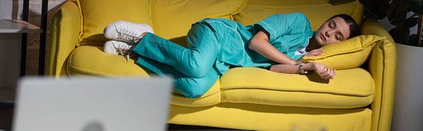 panoramic shot of attractive nurse in uniform sleeping on yellow sofa during night shift - Valokuva, kuva