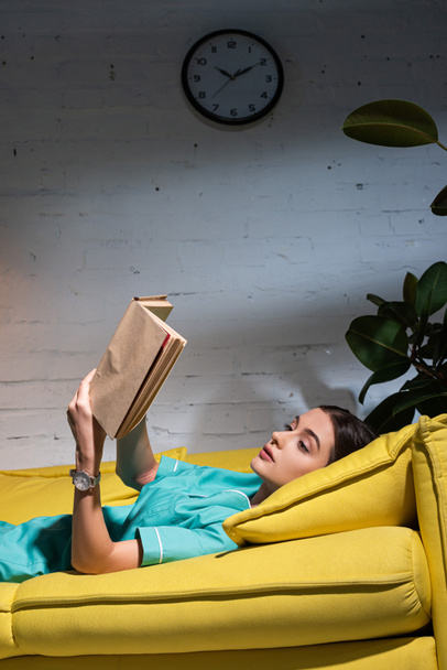 attractive nurse in uniform lying on sofa and reading book during night shift - Fotó, kép