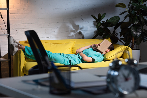 селективное внимание медсестры в форме лежащей на диване с книгой на лице в ночную смену
 - Фото, изображение