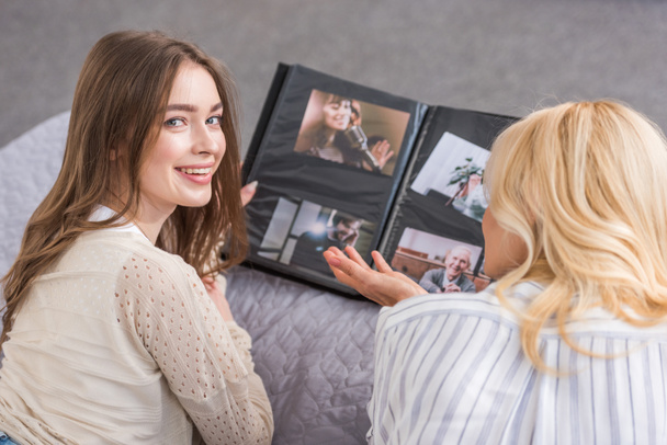 χαρούμενο κορίτσι κοιτάζοντας κάμερα, ενώ βρίσκεται στο κρεβάτι κοντά στη μητέρα κοιτάζοντας άλμπουμ φωτογραφιών - Φωτογραφία, εικόνα