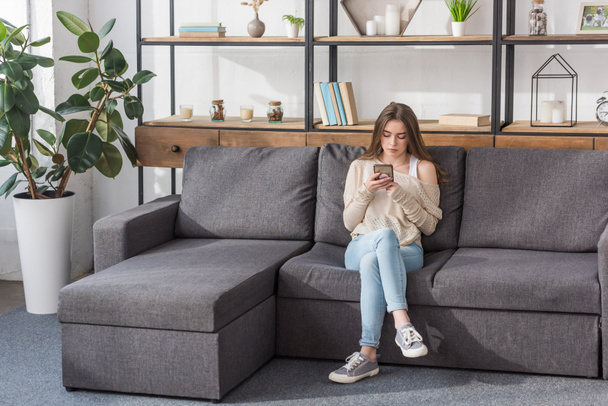 chica bonita usando teléfono inteligente mientras está sentado en el sofá en la sala de estar
 - Foto, Imagen