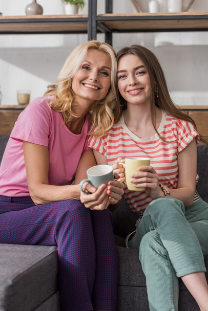 happy mother and daughter looking at camera while sitting on sofa and holding tea cups - Foto, Bild