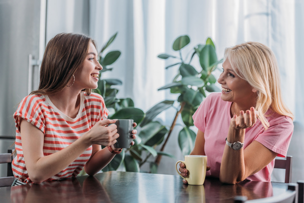 веселая дочь и мать сидят за кухонным столом, разговаривают и пьют чай
 - Фото, изображение