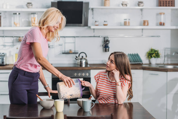 attractive mature woman adding flakes in bowl while young daughter sitting at kitchen table - Foto, immagini