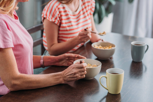 καλλιεργημένη θέα της μητέρας και της κόρης που κάθονται στο τραπέζι της κουζίνας και παίρνουν πρωινό - Φωτογραφία, εικόνα