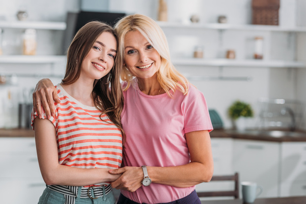 happy mother and daughter hugging and holding hands while smiling at camera in kitchen - Foto, imagen