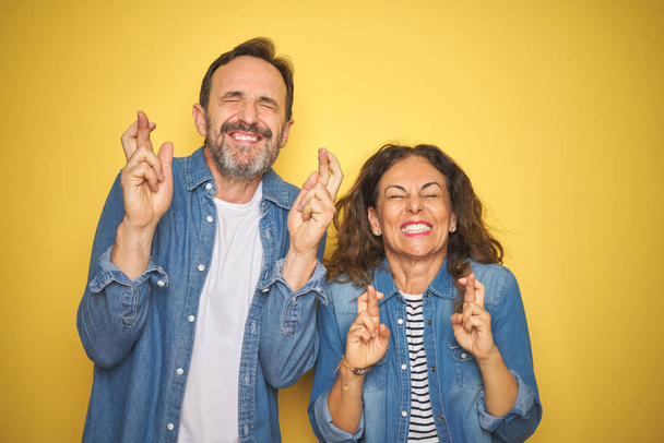 Bella coppia di mezza età insieme indossando camicia di jeans su sfondo giallo isolato gesticolando dito incrociato sorridente con speranza e gli occhi chiusi. Fortuna e concetto superstizioso
. - Foto, immagini