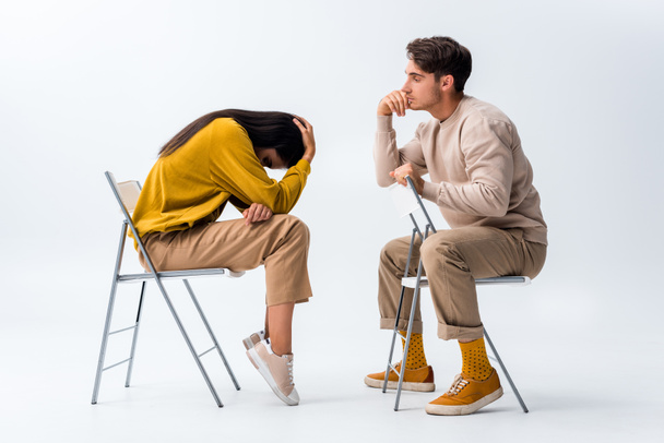 muž sedí na židli a dívá se na rozrušenou ženu zakrývající tvář na bílém  - Fotografie, Obrázek