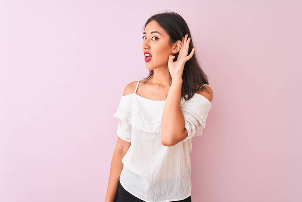 Прекрасна жінка в білій футболці, що стоїть над рожевим фоном, посміхається рукою над вухом, слухаючи слух до чуток або пліток. Концепція глухоти. - Фото, зображення