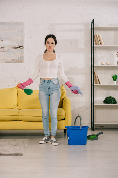young, attractive housewife holding rag and spray bottle while standing near yellow sofa near bucket, brush and scoop on floor - Foto, Imagen