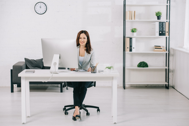 ελκυστική, κομψή γραμματέας χαμογελά στην κάμερα, ενώ κάθεται στο χώρο εργασίας - Φωτογραφία, εικόνα