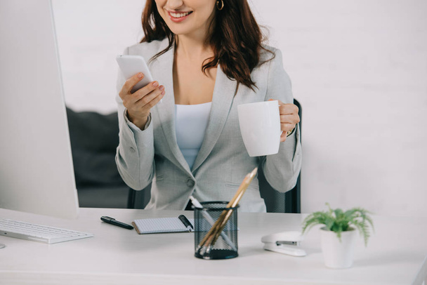 cropped view of smiling secretary holding coffee cup and using smartphone while sitting at workplace - Фото, изображение