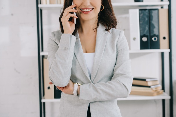 przycięty widok uśmiechniętej, eleganckiej sekretarki rozmawiającej na smartfonie w biurze - Zdjęcie, obraz
