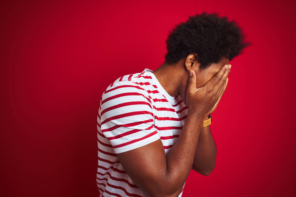 Jovem americano africano com cabelo afro vestindo camiseta listrada sobre fundo vermelho isolado com expressão triste cobrindo o rosto com as mãos enquanto chora. Conceito de depressão
. - Foto, Imagem