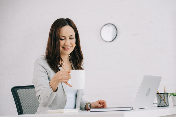 pretty, smiling secretary holding coffee cup while sitting at workplace in office - Φωτογραφία, εικόνα