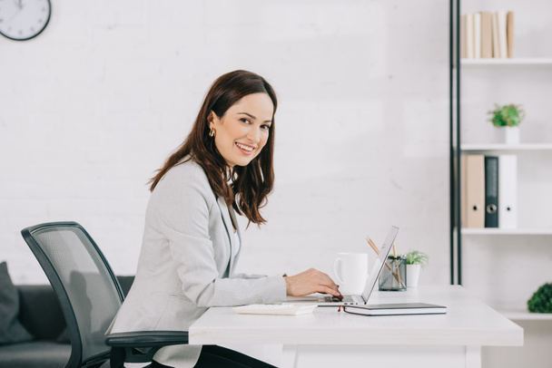 молодая, улыбающаяся секретарша смотрит в камеру, сидя на рабочем месте в офисе
 - Фото, изображение