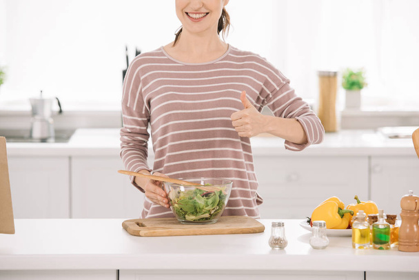 przycięty widok uśmiechniętej kobiety pokazuje kciuk w górę stojąc w pobliżu miski ze świeżą sałatką warzywną  - Zdjęcie, obraz