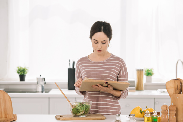 attractive, young woman reading receipt book while standing at kitchen table near bowl with vegetable salad - Photo, Image