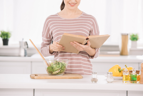 vista recortada de la mujer sonriente lectura libro de recibos, mientras que de pie en la mesa de la cocina cerca de cuenco con ensalada de verduras
 - Foto, imagen
