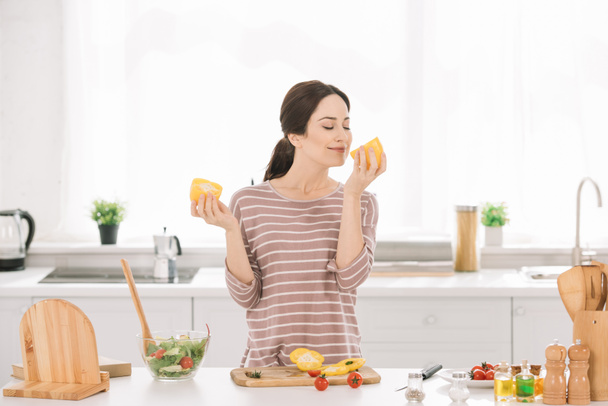 bonita, mujer joven oliendo pimiento cortado mientras está de pie en la mesa de la cocina cerca de verduras frescas
 - Foto, Imagen
