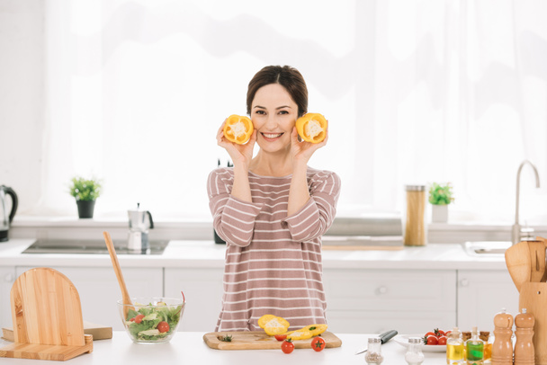 attractive young woman looking at camera while standing near kitchen table with fresh vegetables and holding cut bell pepper  - Fotografie, Obrázek