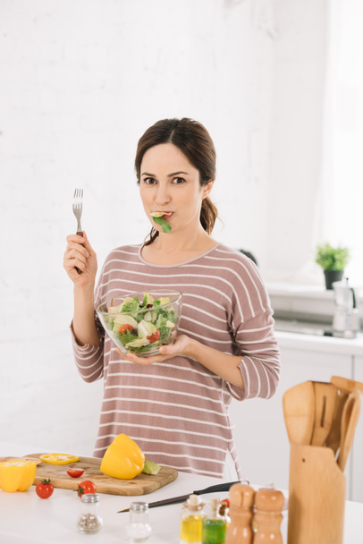 beautiful young woman looking at camera while holding fork and eating fresh vegetable salad  - Fotografie, Obrázek
