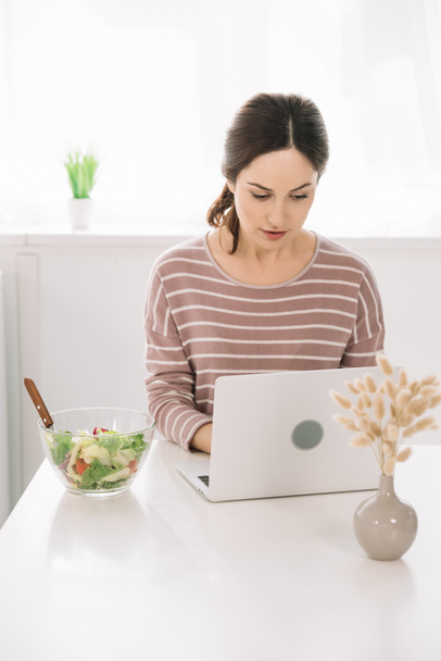 atraktivní, pozorná žena pomocí notebooku při posezení u kuchyňského stolu v blízkosti mísy se zeleninovým salátem - Fotografie, Obrázek