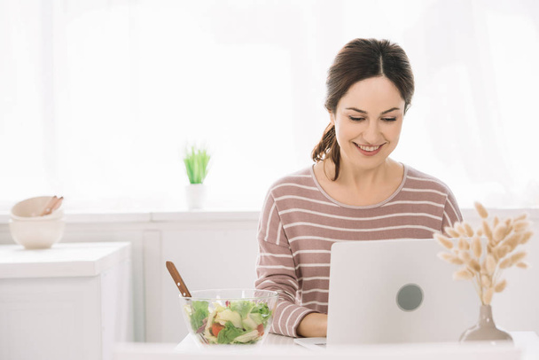 young, happy woman using laptop while sitting at kitchen table near bowl with vegetable salad - 写真・画像