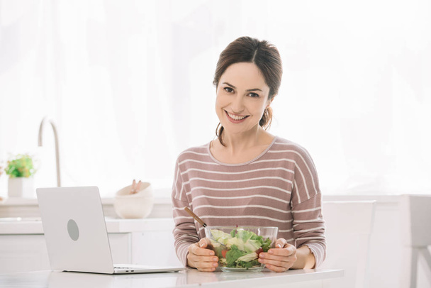 young, smiling woman looking at camera while sitting at table with bowl of vegetable salad near laptop - Photo, image