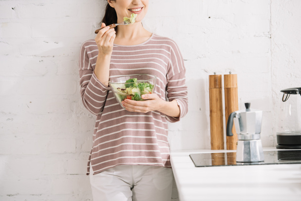 καλλιεργημένη άποψη του χαμογελαστού γυναίκα στέκεται κοντά στον τοίχο και τρώει σαλάτα λαχανικών - Φωτογραφία, εικόνα