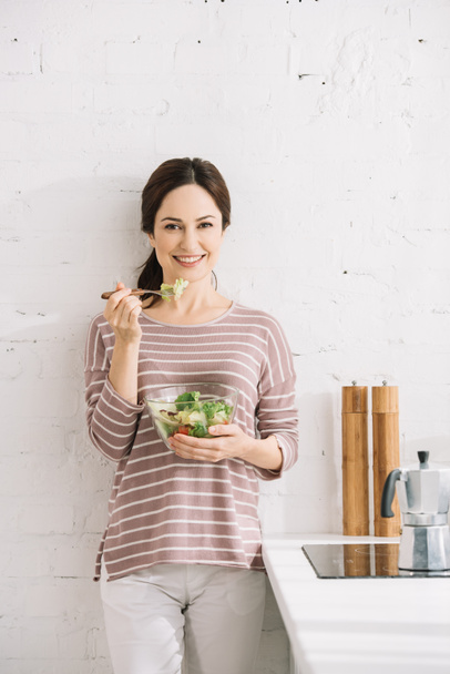 joven, mujer feliz mirando a la cámara mientras está de pie cerca de la pared y comer ensalada de verduras
 - Foto, Imagen