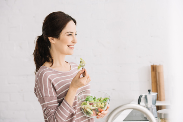 молодая, веселая женщина смотрит в сторону, когда ест овощной салат
 - Фото, изображение