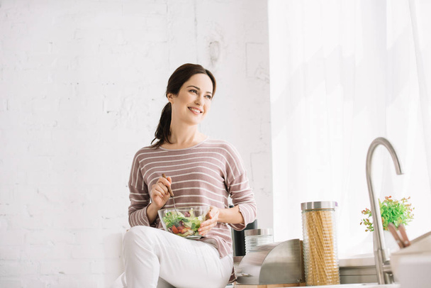 счастливая молодая женщина смотрит в сторону, сидя на кухонном столе и держа миску с овощным салатом
 - Фото, изображение